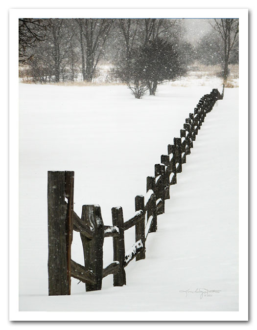 Falling snow blankets a split rail fence with trees in the background.
